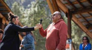 woman hands microphone to man in red shirt outside under wooden roof with trees in background