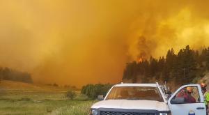 A BC government truck and wildfire personnel on a road with a wall of smoke and fire in the background