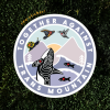 Circle-shaped sticker on a mossy green background. Sticker says "Together Against Trans Mountain" and features artwork of 5 animal species